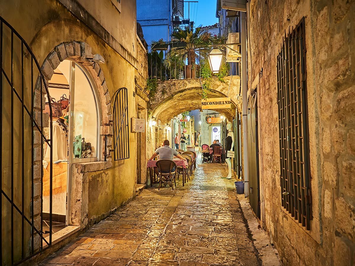 Beleuchtete historische Gasse mit Steinhäusern, Restaurants und Geschäften am Abend in einer mediterranen Altstadt.