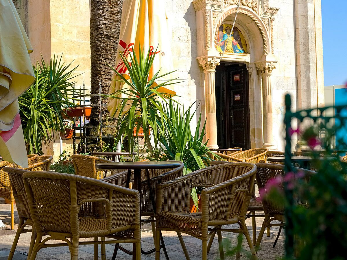 Sonniges Straßencafé mit Korbsesseln und Tischen vor einer historischen Kirche mit Mosaikportal, Palmen und grünen Pflanzen.