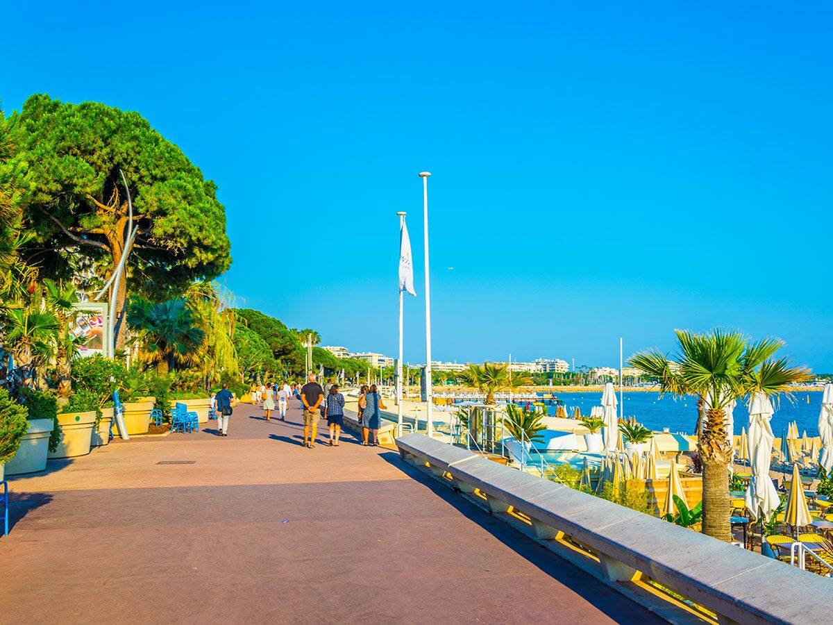 Menschen spazieren auf der sonnigen Promenade von Cannes entlang des Mittelmeeres mit Strand und Palmen unter blauem Himmel.