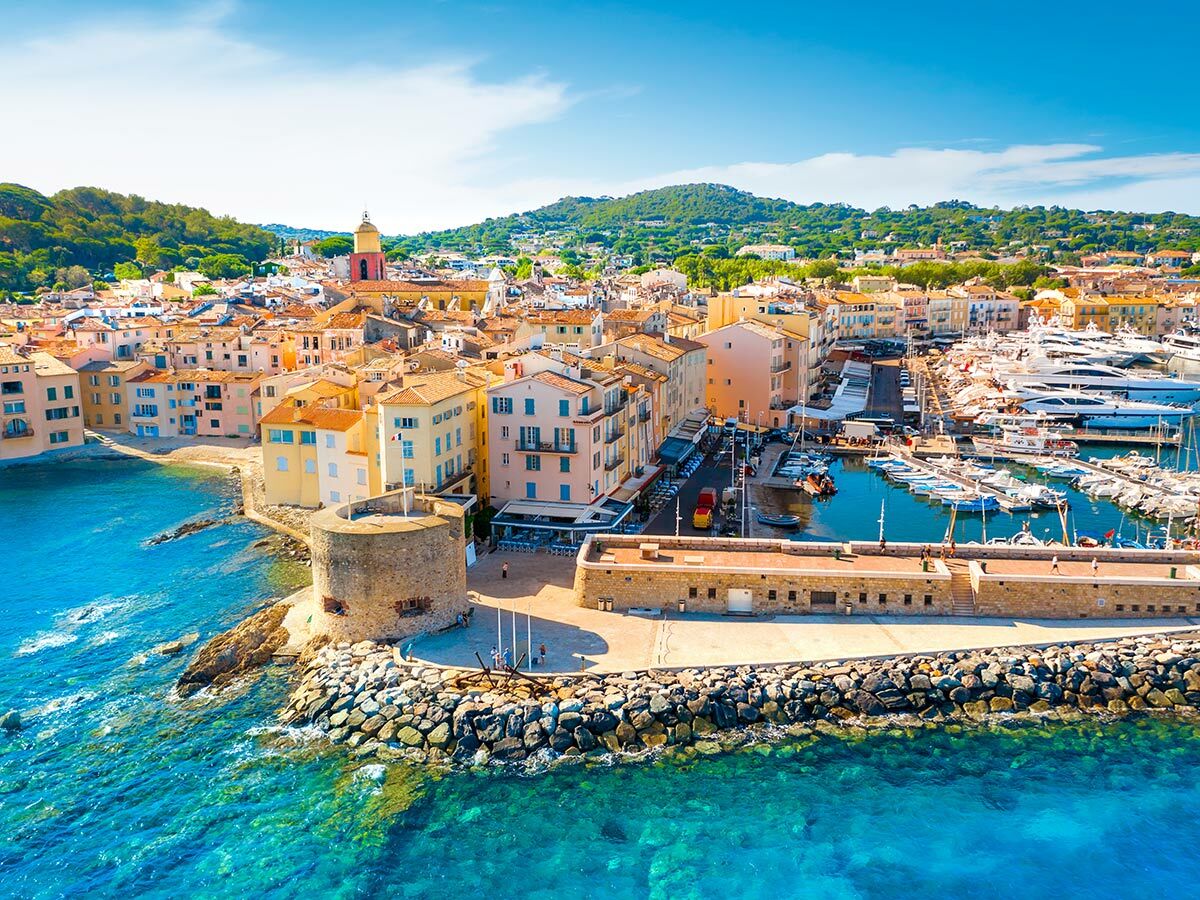 Luftaufnahme von Saint-Tropez mit farbenfrohen Häusern, Yachthafen, Kirchturm und türkisblauem Mittelmeer an einem sonnigen Tag.