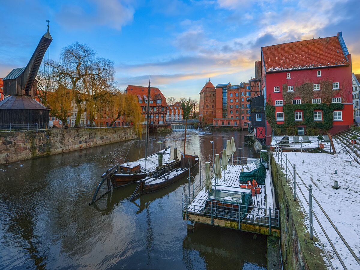 Historische Altstadt von Lüneburg im Winter mit dem Alten Kran, verschneiten Booten und roten Gebäuden am Flussufer unter farbigem Himmel.