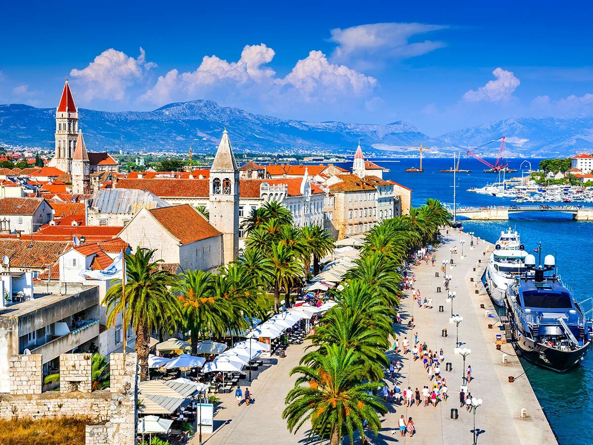 Blick auf die historische Stadt Trogir, Kroatien, mit Kathedrale, roten Dächern, belebter Promenade, Palmen und Yachthafen am Adriatischen Meer.