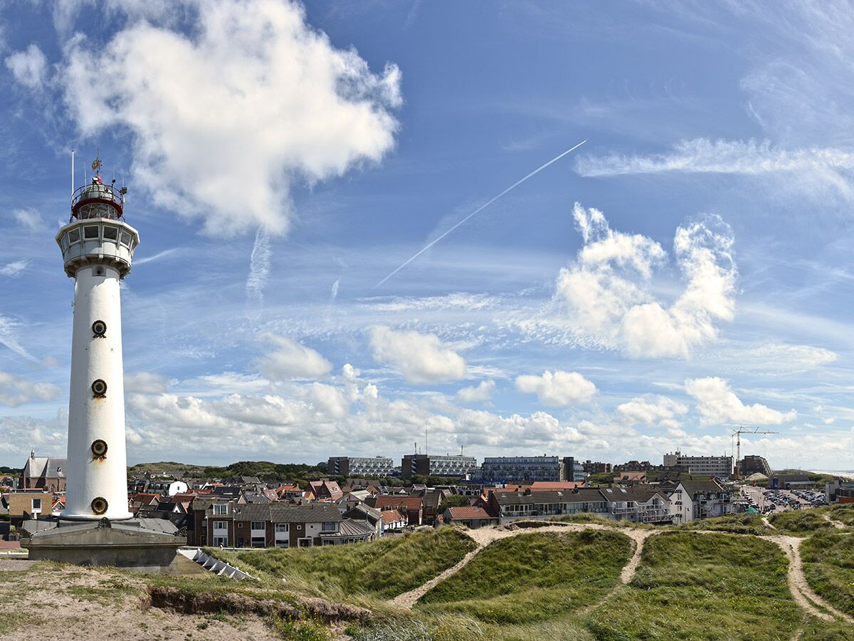 Hoher weißer Leuchtturm vor blauem Himmel mit Wolken und einem Küstenort mit Häusern und Dünen im Hintergrund.