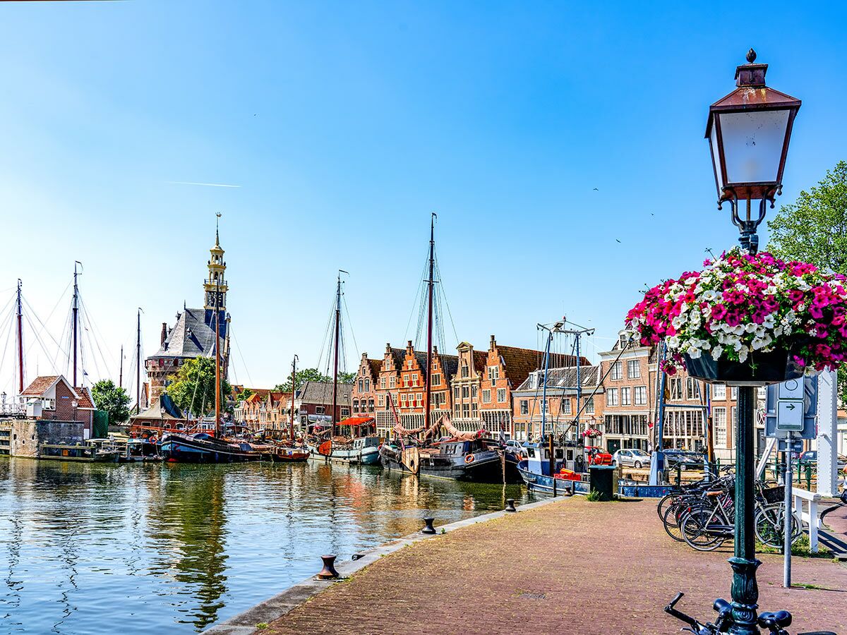 Historischer Hafen Niederlande: Segelboote, Giebelhäuser, Turm, gepflasterte Promenade, Blumen, Fahrräder unter blauem Himmel am Wasser.