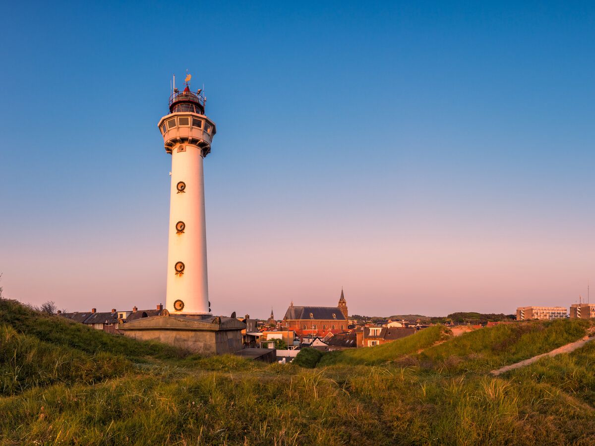 Weißer Leuchtturm in Egmond aan Zee bei Sonnenuntergang über grünen Dünen und Küstenstadt.