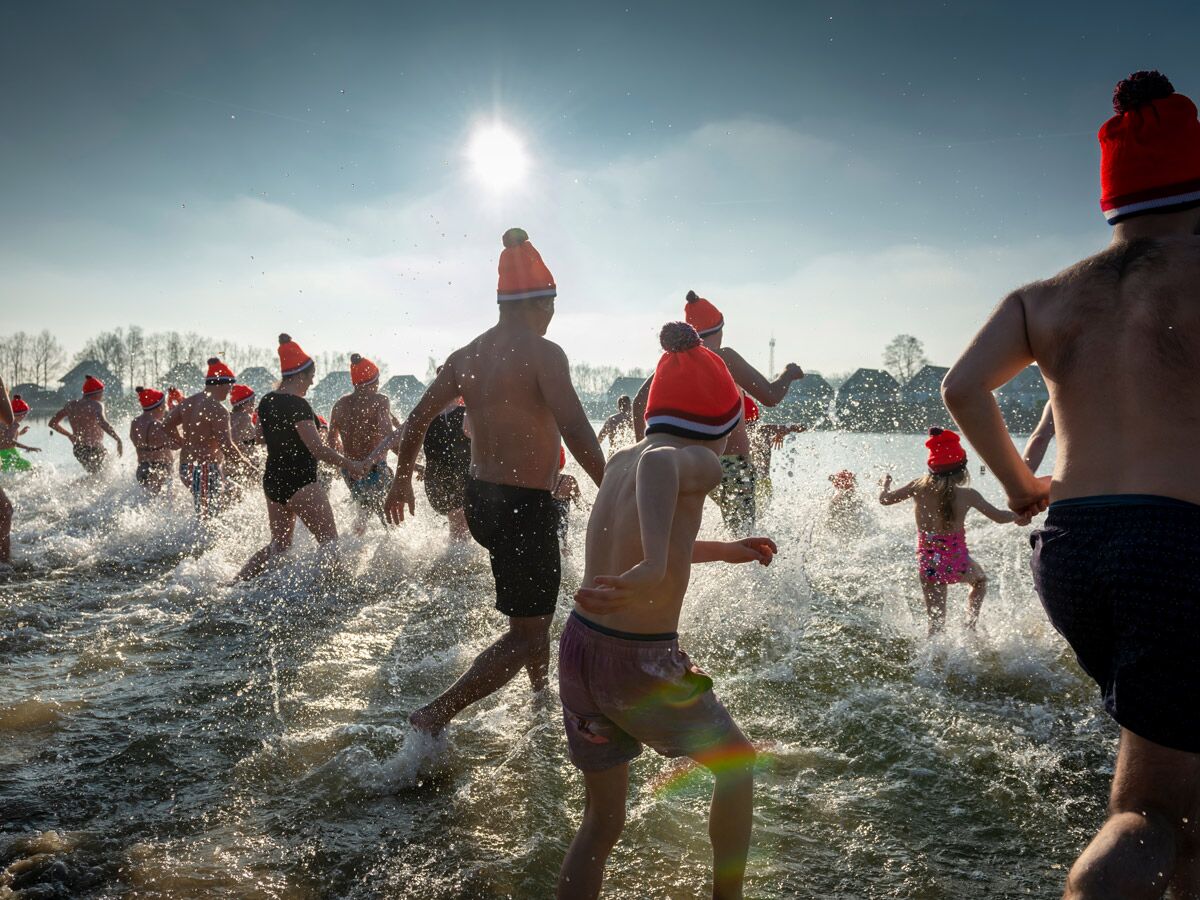 Menschen mit orangefarbenen Mützen rennen unter strahlender Sonne ins kalte Wasser, spritzen Wasser für Neujahrstauchen.