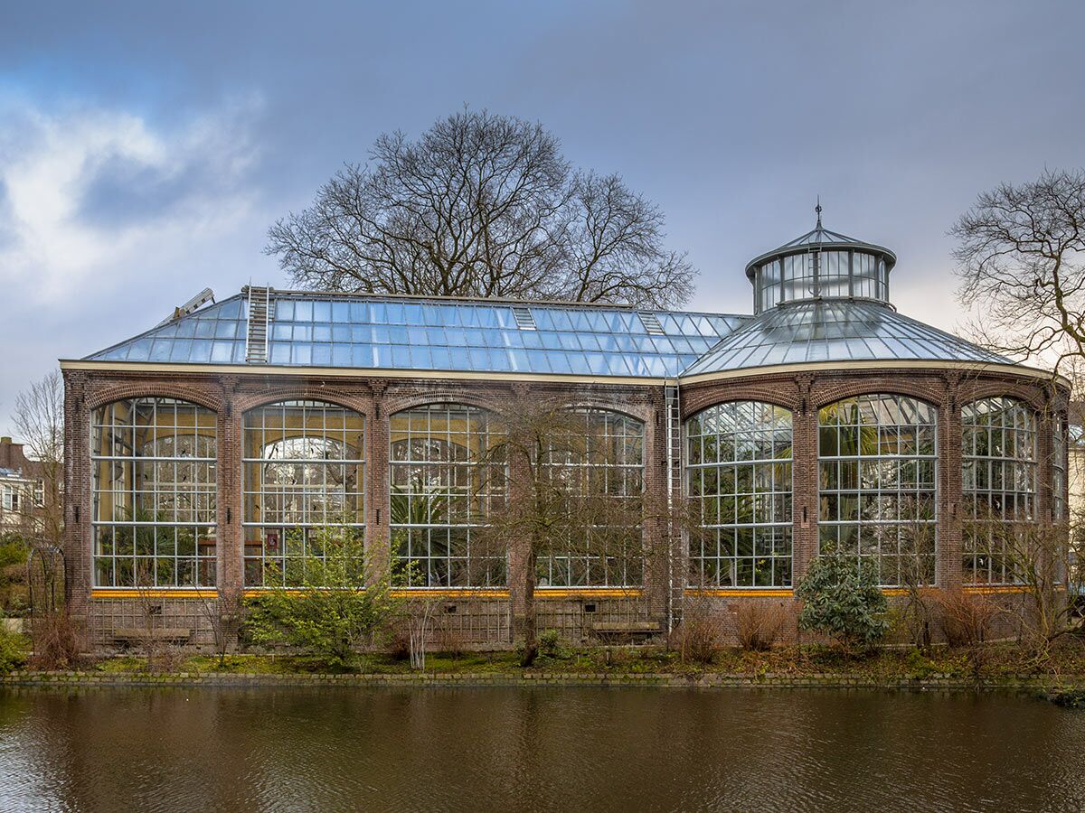 Historisches Backstein-Gewächshaus mit Glasdach und großen Fenstern, gespiegelt in einem Gewässer, unter bewölktem Himmel.
