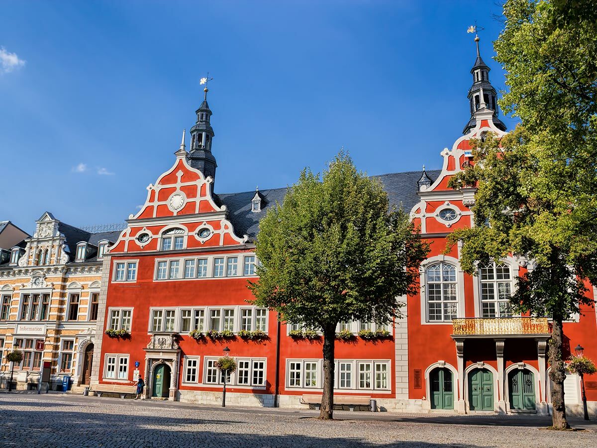 Rotes, historisches Rathaus mit zwei Giebeln, Türmen, weißen Verzierungen und einem hellen Gebäude auf sonnigem Marktplatz.