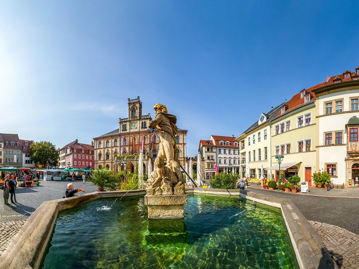Sonnenbeschienener historischer Marktplatz mit barockem Neptunbrunnen, geschäftigen Menschen und farbigen Häusern sowie einem Rathaus.