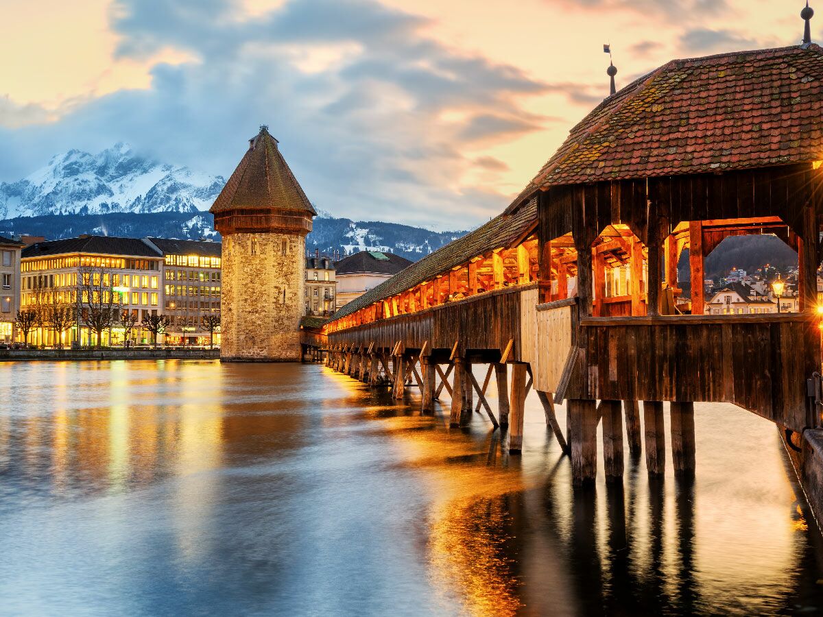 Historische Kapellbrücke und Wasserturm in Luzern, Schweiz, bei Sonnenuntergang beleuchtet. Stadt und schneebedeckte Berge im Hintergrund.