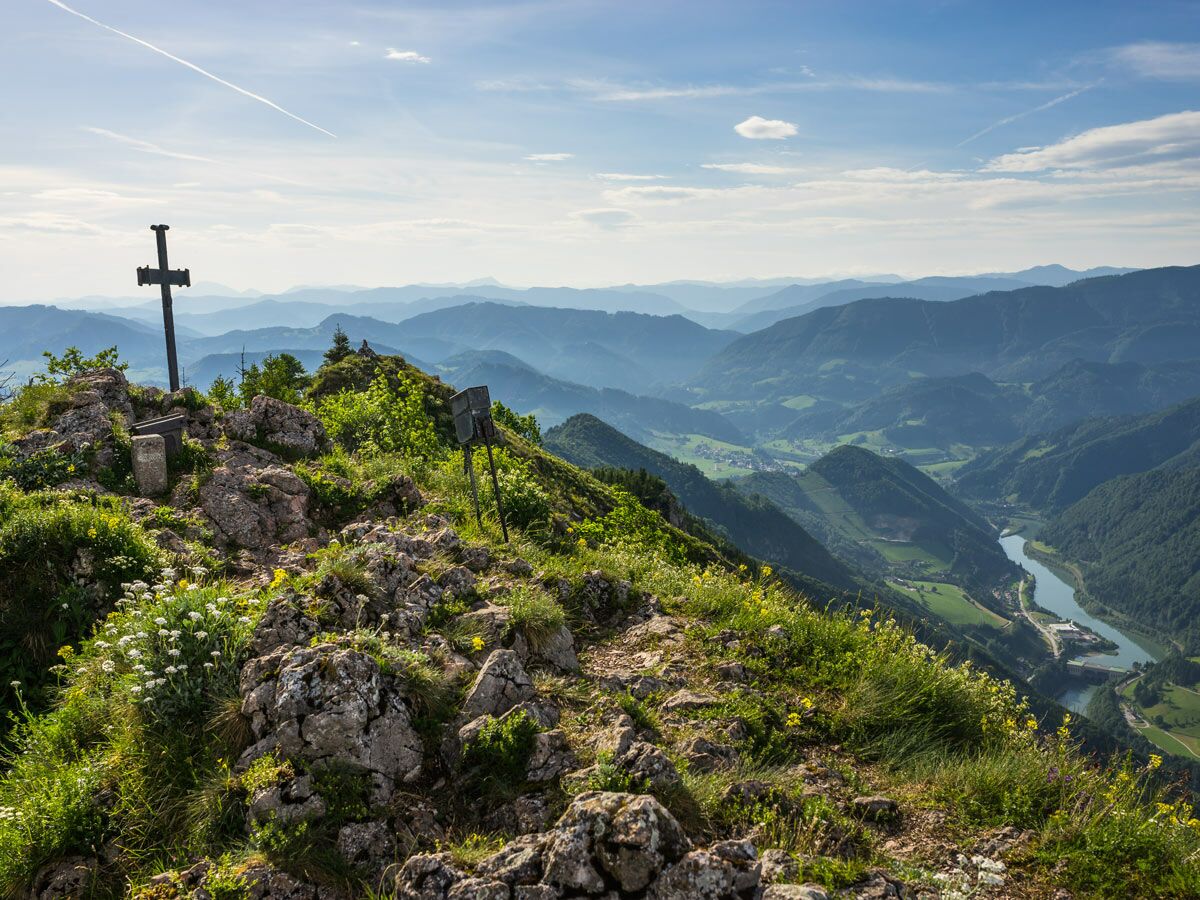 Gipfelkreuz auf felsigem Berg mit grünen Wiesen, weiten blauen Bergen, einem Fluss im Tal und klarem Himmel.
