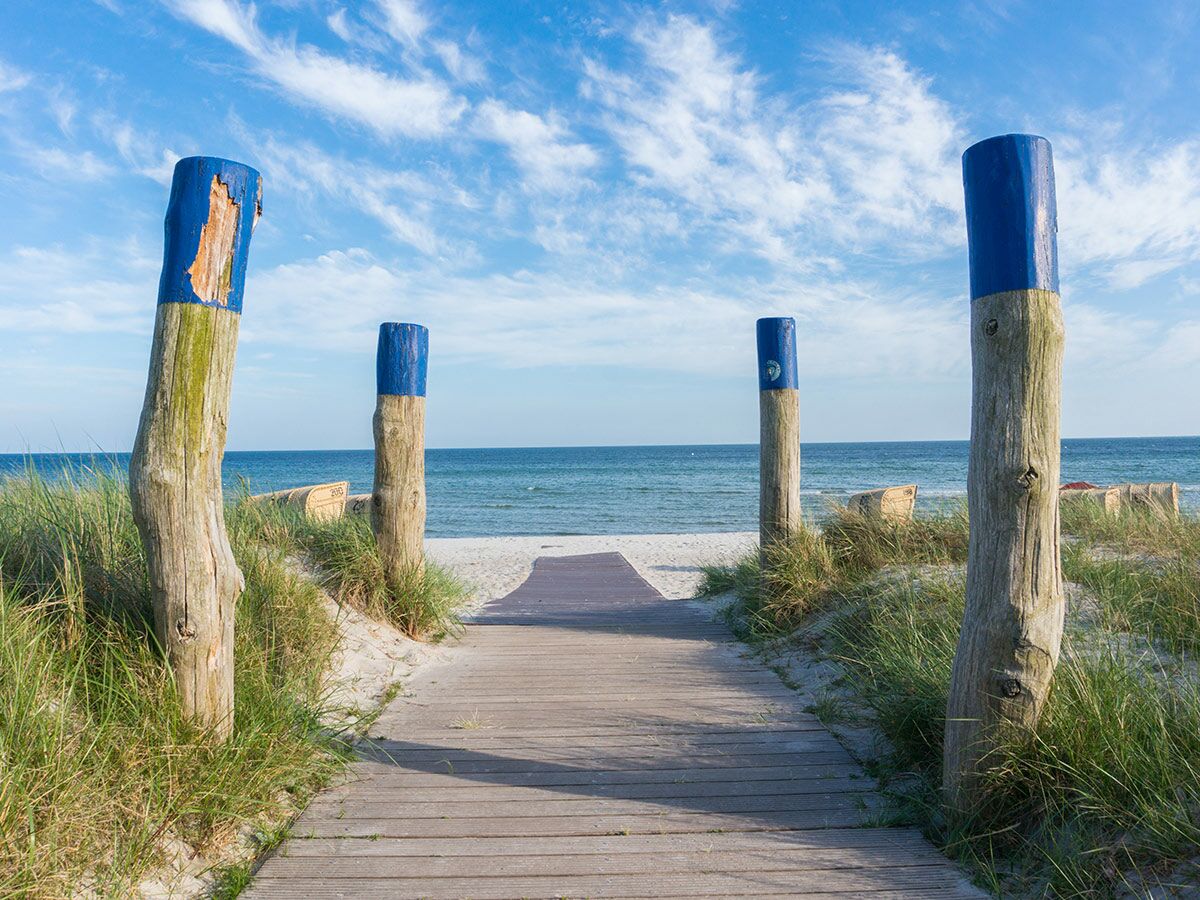 Ein Holzsteg führt durch Sanddünen mit blauen Pfosten zu einem sonnigen Sandstrand und dem blauen Meer.