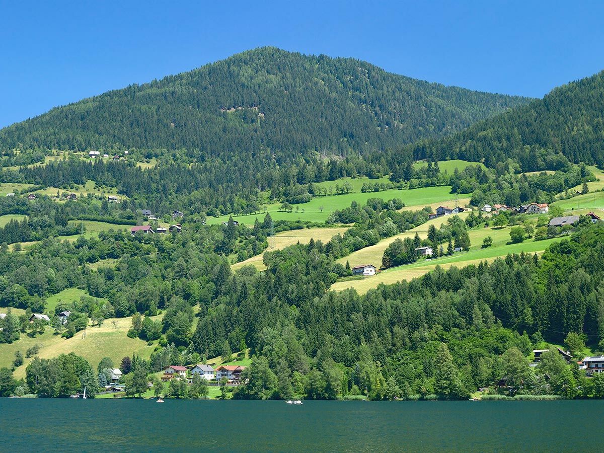 Idyllische Alpenlandschaft mit einem See im Vordergrund, grünen bewaldeten Bergen und einem Dorf am Hang unter blauem Himmel.