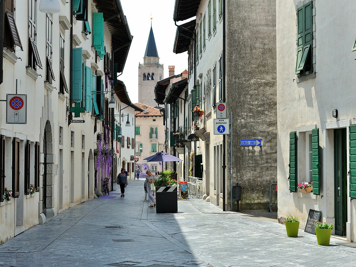 Schmale Gasse in italienischer Altstadt mit weißen Gebäuden, grünen Fensterläden, Menschen und einem Kirchturm im Hintergrund.