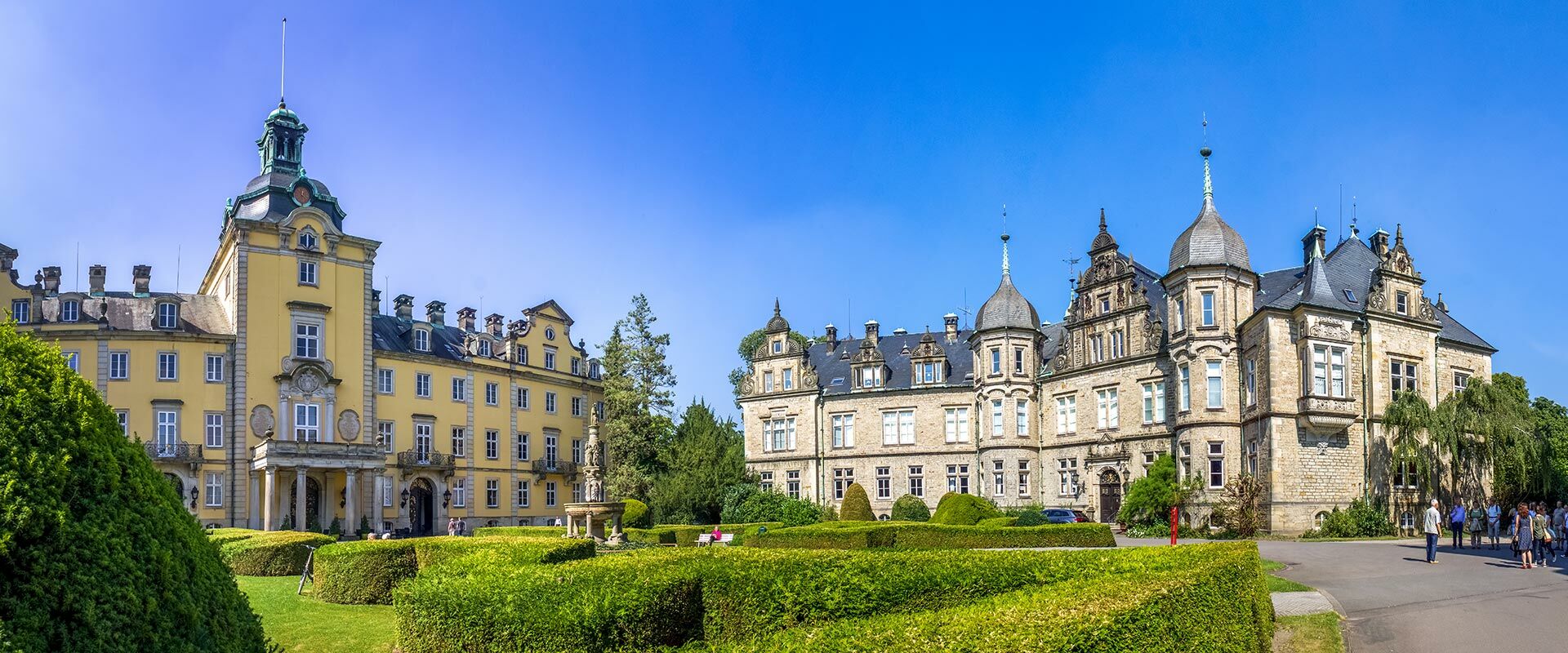 Historisches Schloss Bückeburg mit gelbem Barockflügel und steinernem Anbau, umgeben von gepflegten Hecken im Schlosspark bei Sonnenschein.