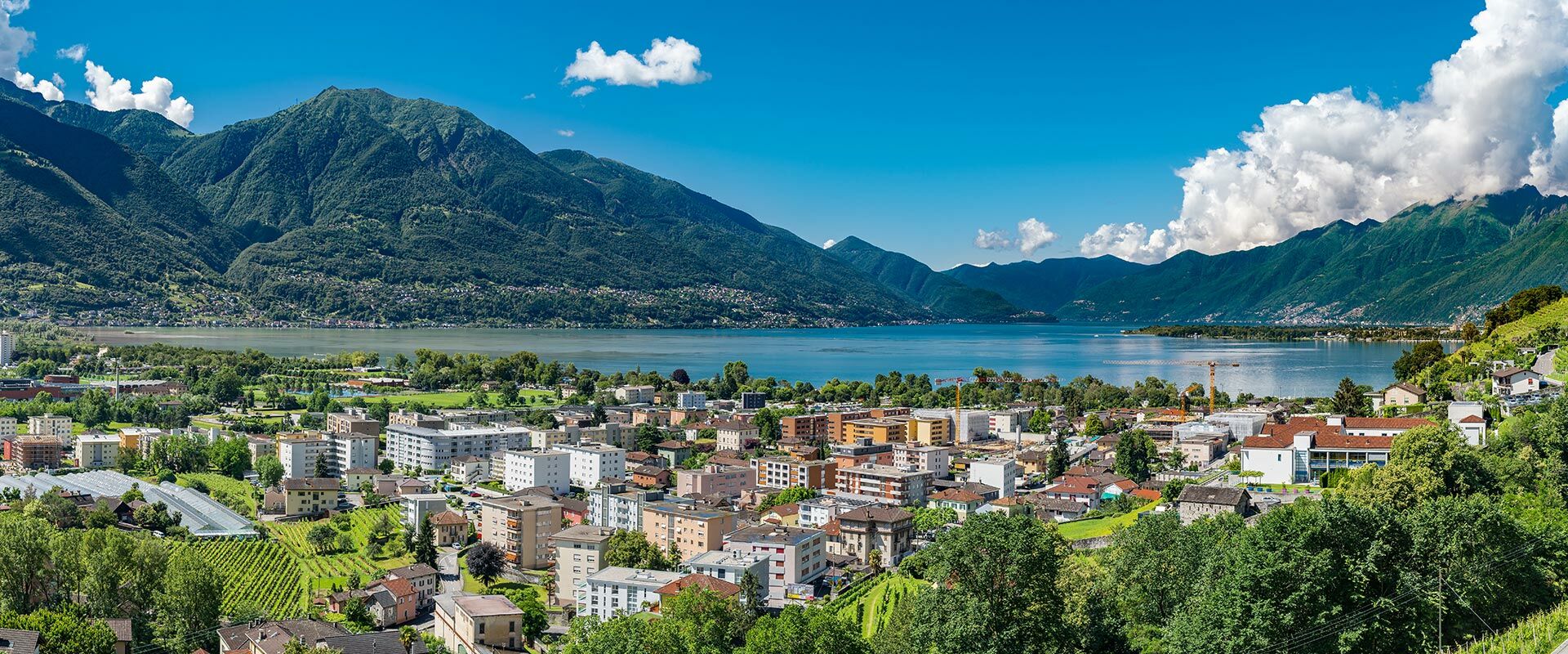 Panoramablick auf eine Stadt am Ufer eines großen Sees, umgeben von grünen Bergen unter blauem Himmel mit Wolken.