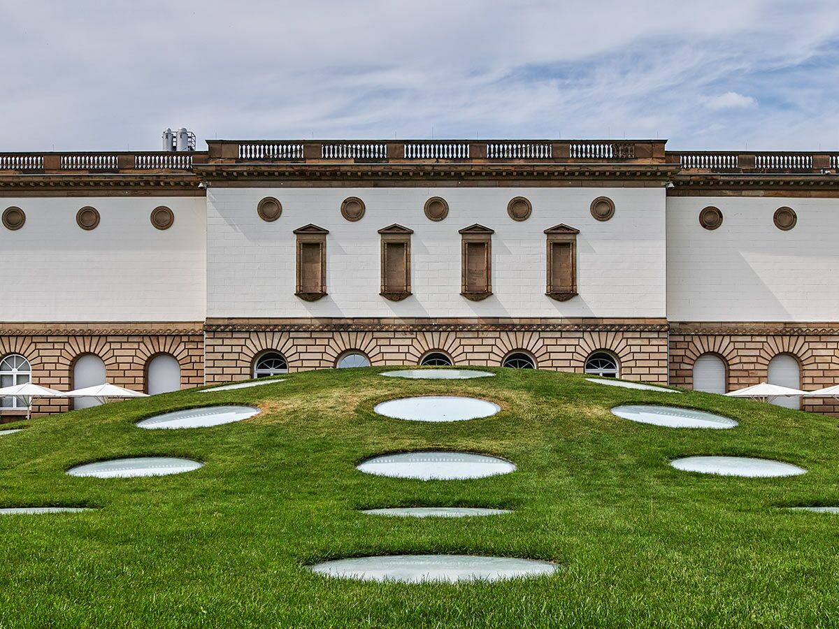 Die Fassade eines klassischen Gebäudes trifft auf einen modernen grünen Rasenhügel mit runden, hellen Lichtkuppeln im Vordergrund.