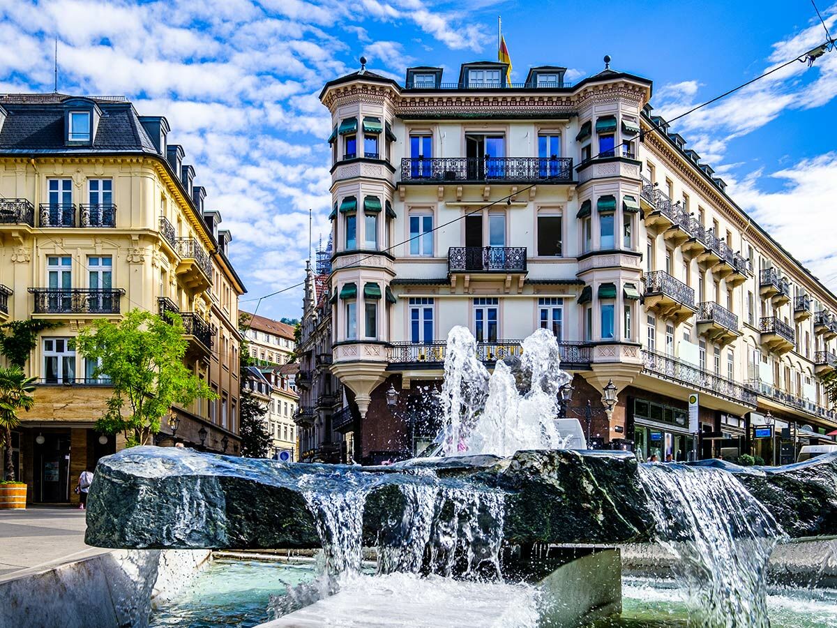 Modernes Stadtbrunnen mit Wasserspielen vor prächtigen historischen Gebäuden in Baden-Baden unter blauem Himmel.
