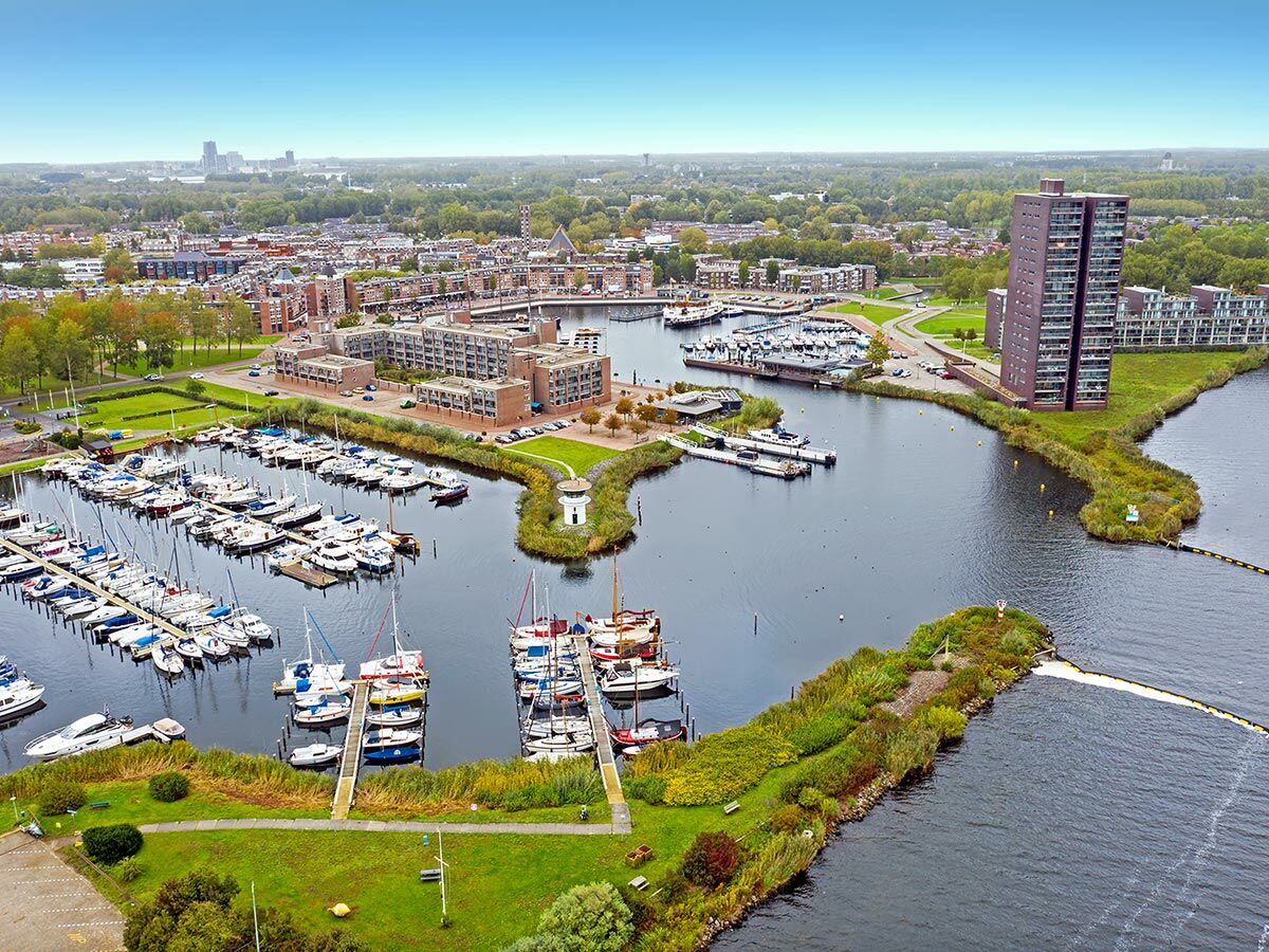 Luftaufnahme belebter Marina mit Segel- und Motorbooten an Stegen in Stadtlandschaft, umgeben von Gebäuden und Grünflächen.