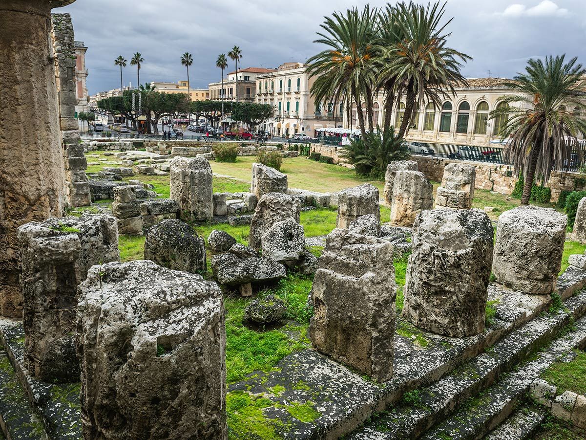 Antike Ruinen des Tempels von Apollo in Syrakus, Sizilien, mit massiven Steinblöcken, moosbewachsenem Gras und Stadt im Hintergrund.