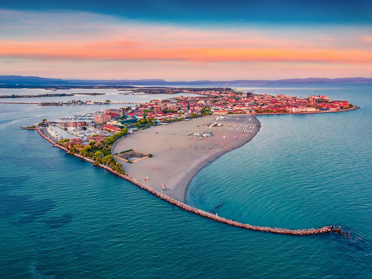 Luftaufnahme einer malerischen Küstenstadt mit Yachthafen, Sandstrand und Wellenbrecher bei farbenprächtigem Sonnenuntergang.