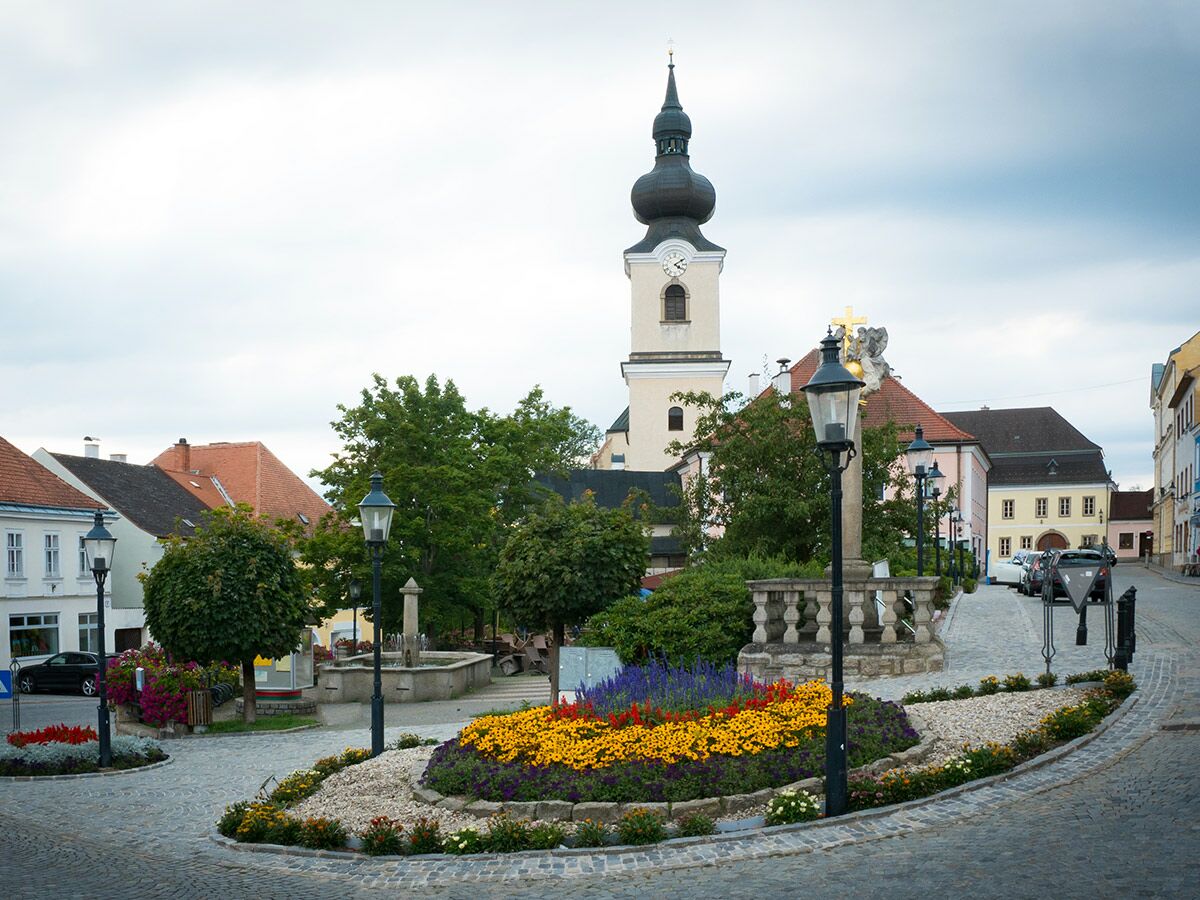 Idyllischer europäischer Stadtplatz mit historischem Kirchturm, farbenfrohen Blumenbeeten, Kopfsteinpflaster, alten Gebäuden und Laternen.