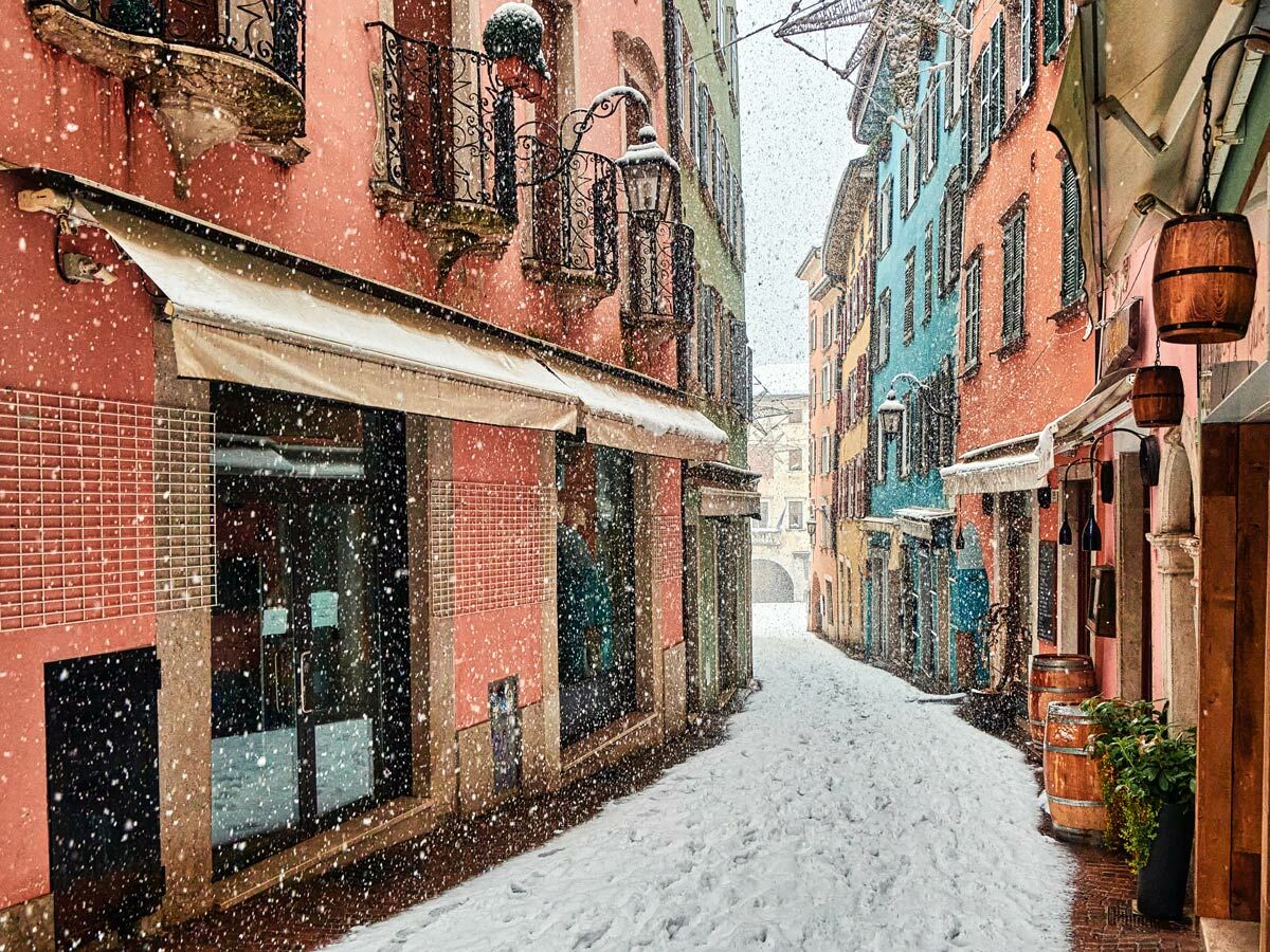 Blick auf eine verschneite, enge Gasse mit farbenfrohen Häusern, Geschäften und Balkonen bei leichtem Schneefall.