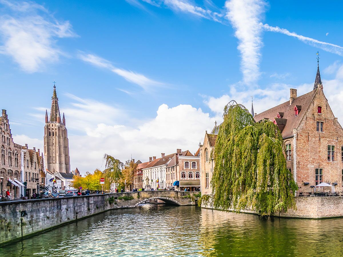 Blick auf den Kanal in Brügge mit alten Backsteinhäusern, dem Turm der Liebfrauenkirche und einem Weidenbaum am Wasser.