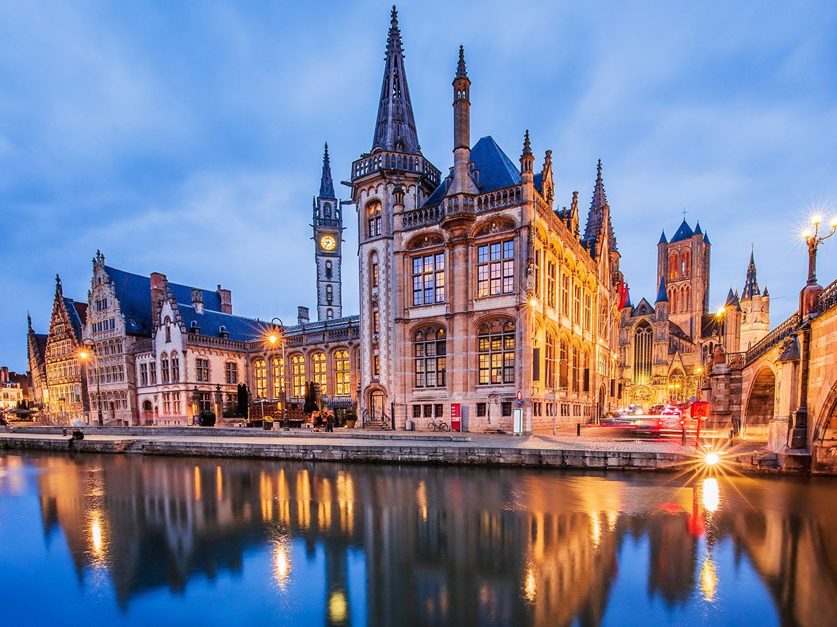 Beleuchtete historische Gebäude der Altstadt von Gent entlang des Kanals Leie bei Nacht mit leuchtenden Spiegelungen im Wasser.
