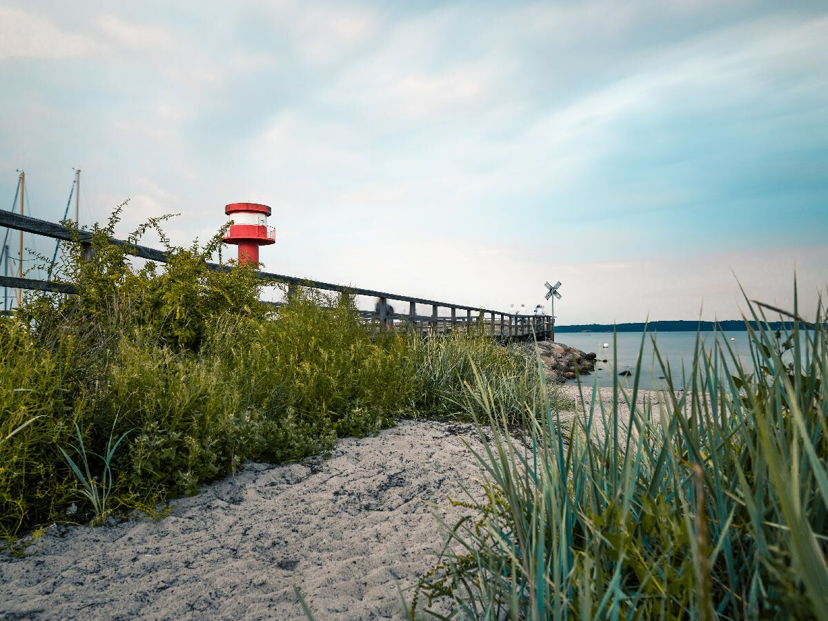 Küstenweg mit Strand, grünen Gräsern und Büschen, hölzernem Pier, rotem Leuchtturm und blauem Meer unter bewölktem Himmel.