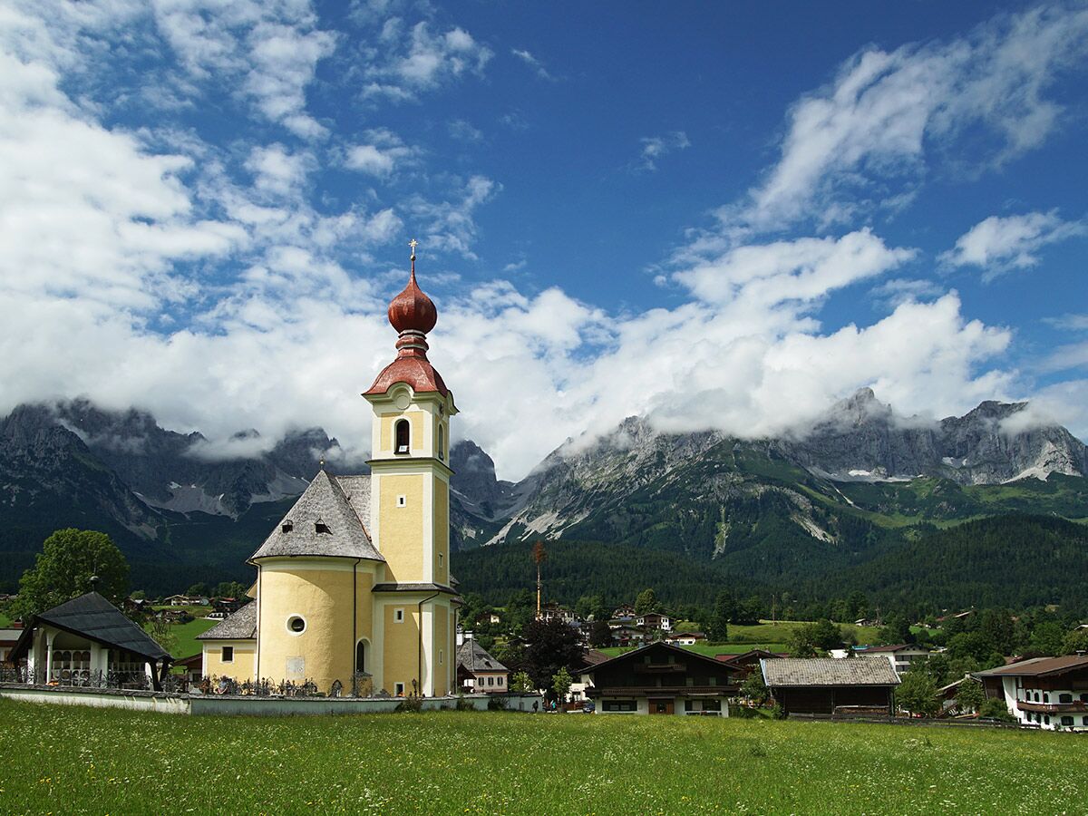 Barocke Kirche mit Zwiebelturm vor majestätischen Alpenbergen und grünen Wiesen unter blauem Himmel mit Wolken.