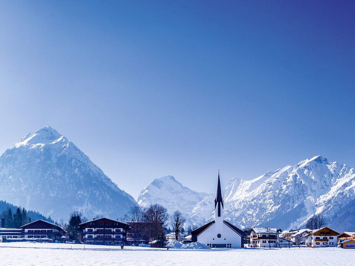 Idyllische Alpen Winterlandschaft mit schneebedeckten Bergen, einem malerischen Dorf und einer weißen Kirche unter blauem Himmel.