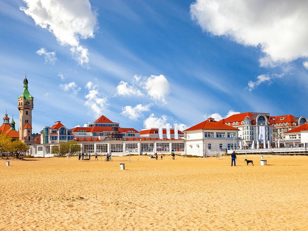 Strand von Sopot mit Leuchtturm, historischen Gebäuden, roten Dächern und Menschen am Meer unter blauem Himmel.