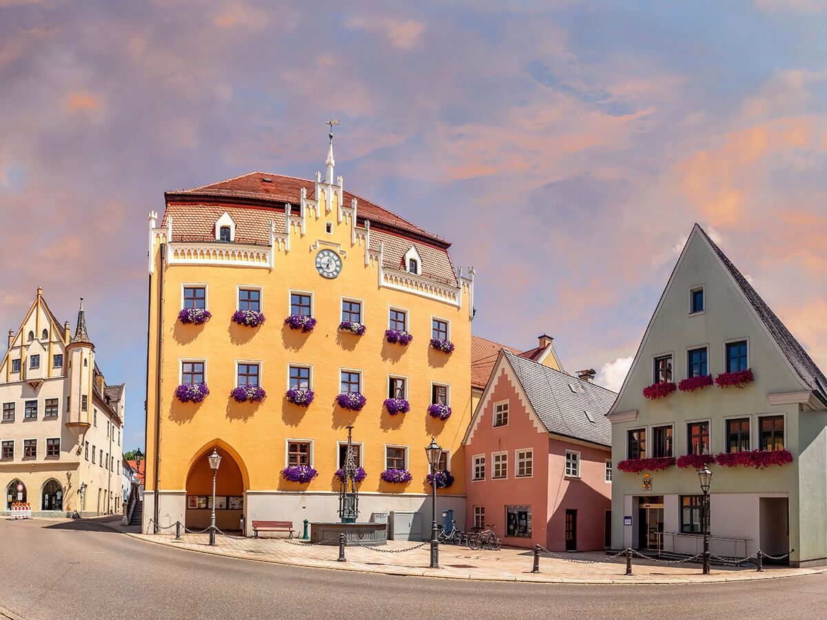 Historische Gebäude in europäischer Altstadt mit gelbem Rathaus, rosa und grünem Haus, Blumenkästen, Brunnen unter farbigem Himmel.