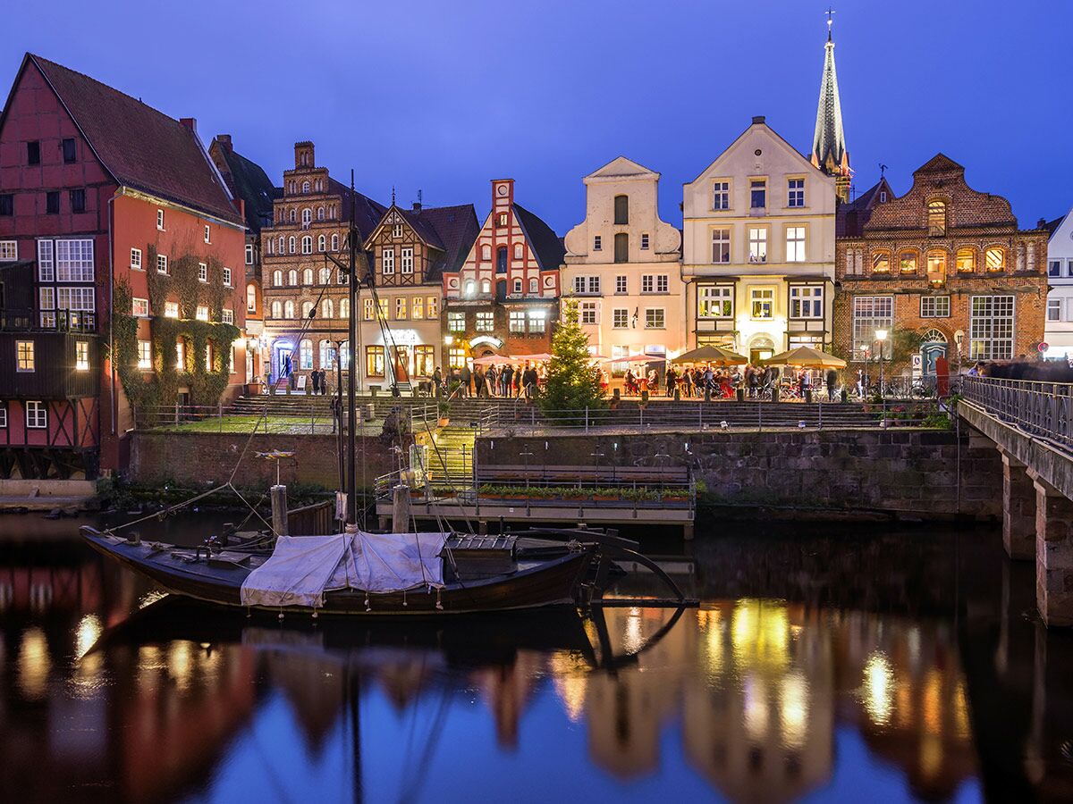 Historische Gebäude am Fluss in Lüneburg bei Abenddämmerung, beleuchtet mit Boot und Spiegelungen im Wasser.