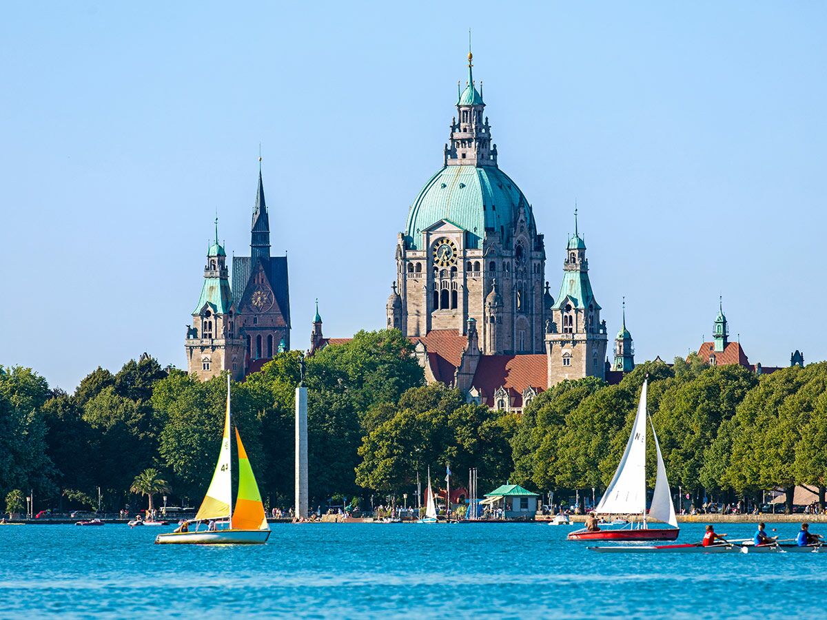 Segelboote und Ruderboote auf dem Maschsee in Hannover mit dem Neuen Rathaus und grünen Bäumen im Hintergrund an einem sonnigen Tag.