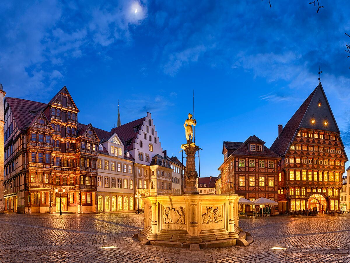 Historischer deutscher Marktplatz bei Dämmerung mit beleuchteten Fachwerkhäusern, Brunnen und Mond am Himmel.