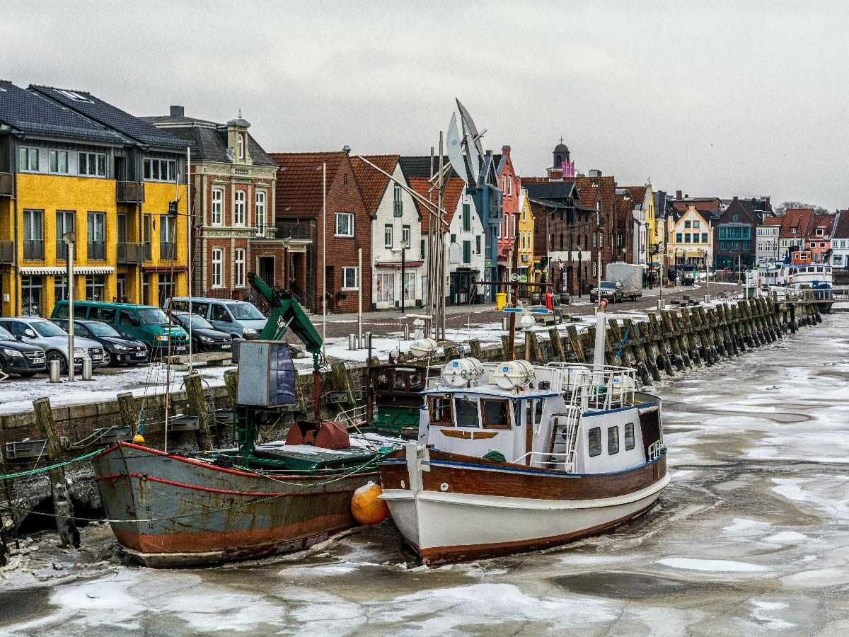 Blick auf einen gefrorenen Hafen mit zwei Booten und bunten Gebäuden an der Uferpromenade bei bewölktem Winterwetter.