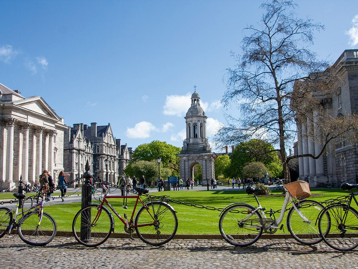 Blick auf den belebten Campus des Trinity College Dublin mit Fahrrädern, historischen Gebäuden und dem Glockenturm an einem sonnigen Tag.