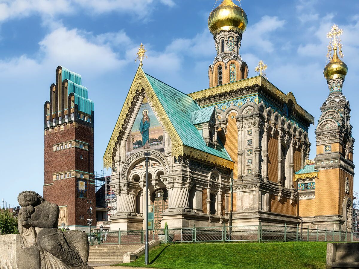 Die Russische Kapelle und der Hochzeitsturm auf der Mathildenhöhe in Darmstadt unter blauem Himmel, mit Skulptur im Vordergrund.