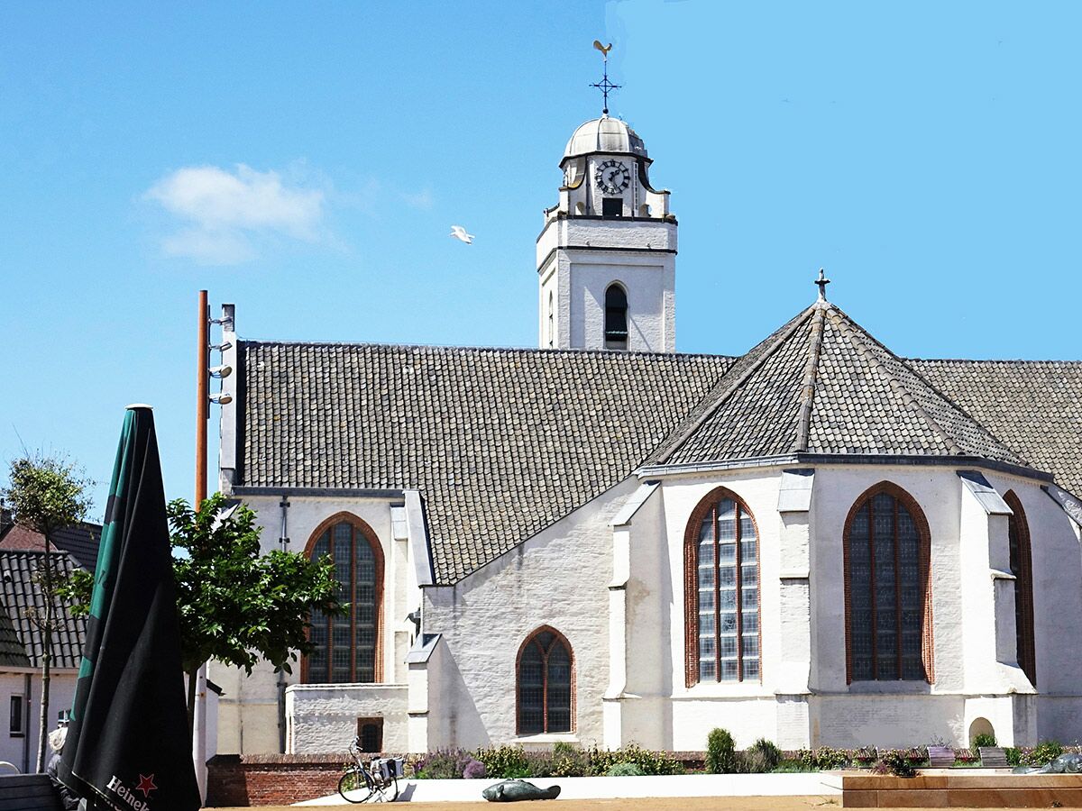 Weiße Kirche mit Glockenturm und Uhr unter blauem Himmel, ein zusammengeklappter Sonnenschirm und Fahrrad im Vordergrund.