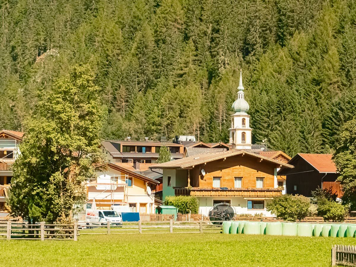 Idyllisches Alpendorf mit traditionellen Holzhäusern, einer Kirche mit Zwiebelturm, Wiesen und Waldhügeln.