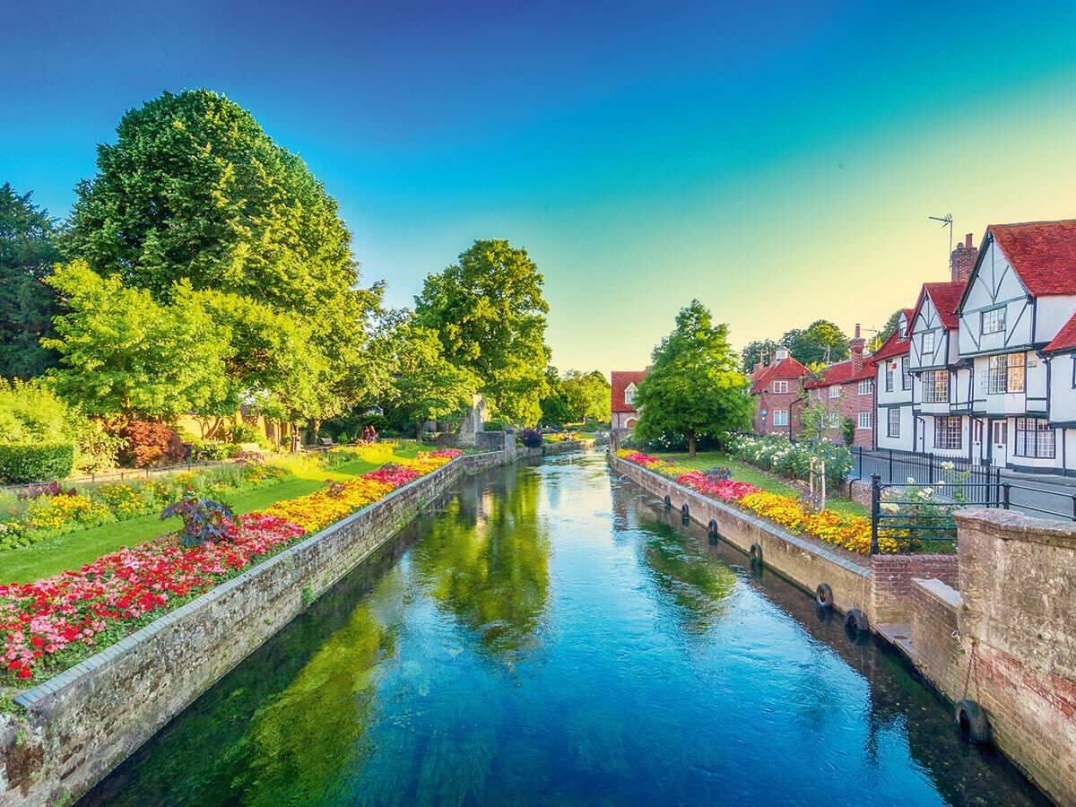 Malerischer Kanal mit bunten Blumenbeeten, grünen Bäumen und traditionellen Fachwerkhäusern unter blauem Himmel.