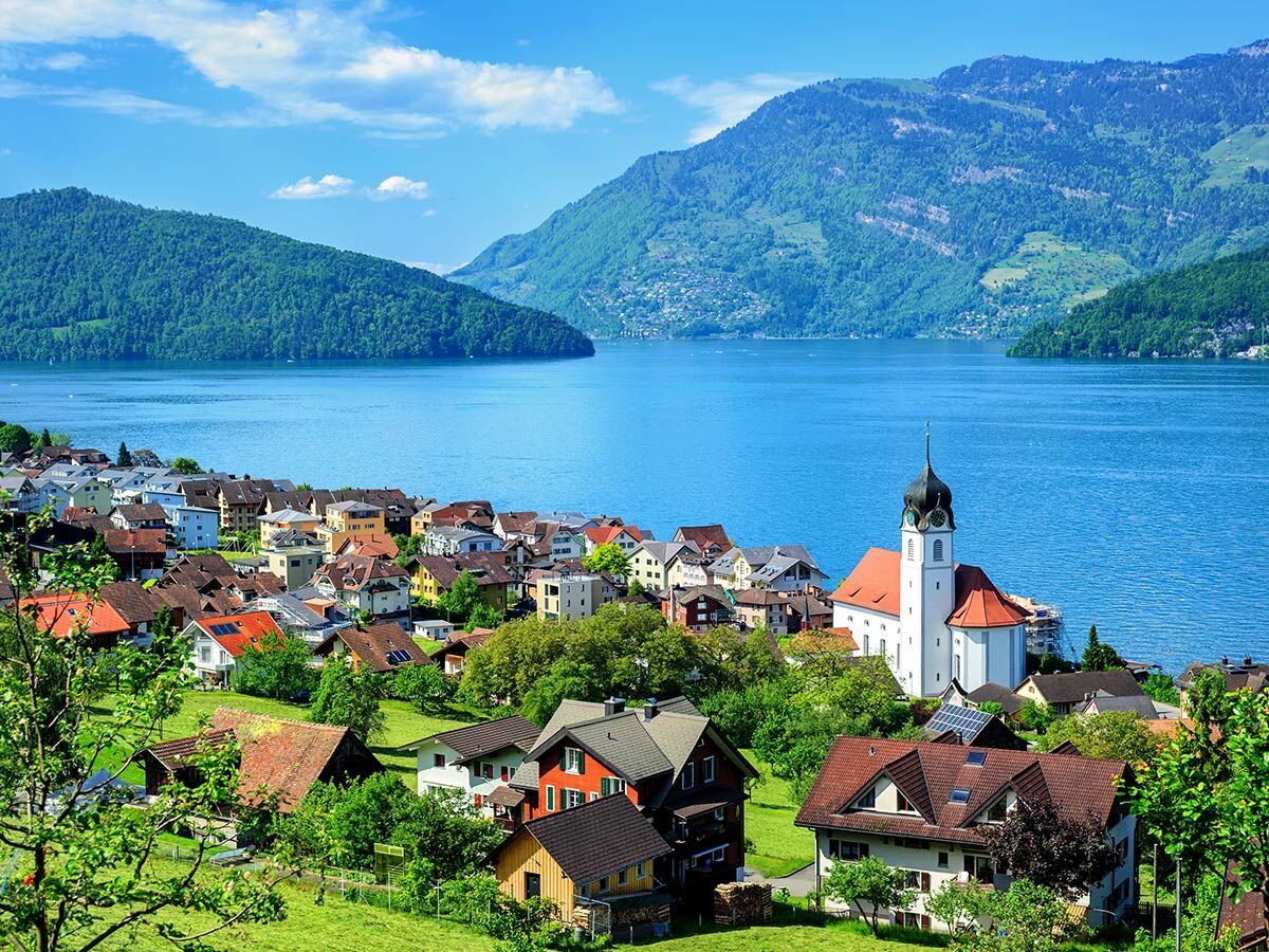 Panoramabild des Schweizer Dorfes Vitznau mit Kirche, Häusern, grünen Hügeln und Bergen am blauen Vierwaldstättersee.