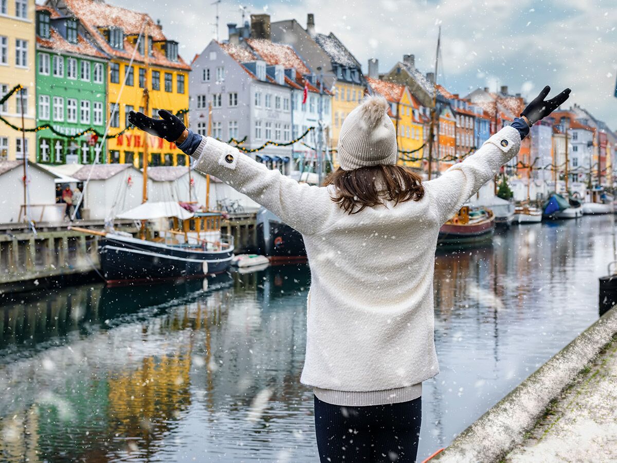 Frau genießt schneebedecktes Nyhavn, Kopenhagen, mit ausgestreckten Armen vor bunten Häusern und Kanal mit Booten.