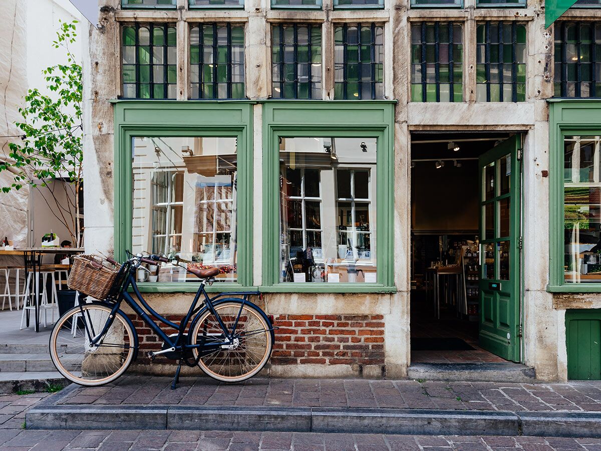 Dunkelblaues Hollandrad mit Weidenkorb vor grünem Laden in europäischer Altstadt. Offene Tür, reflektierende Fenster.