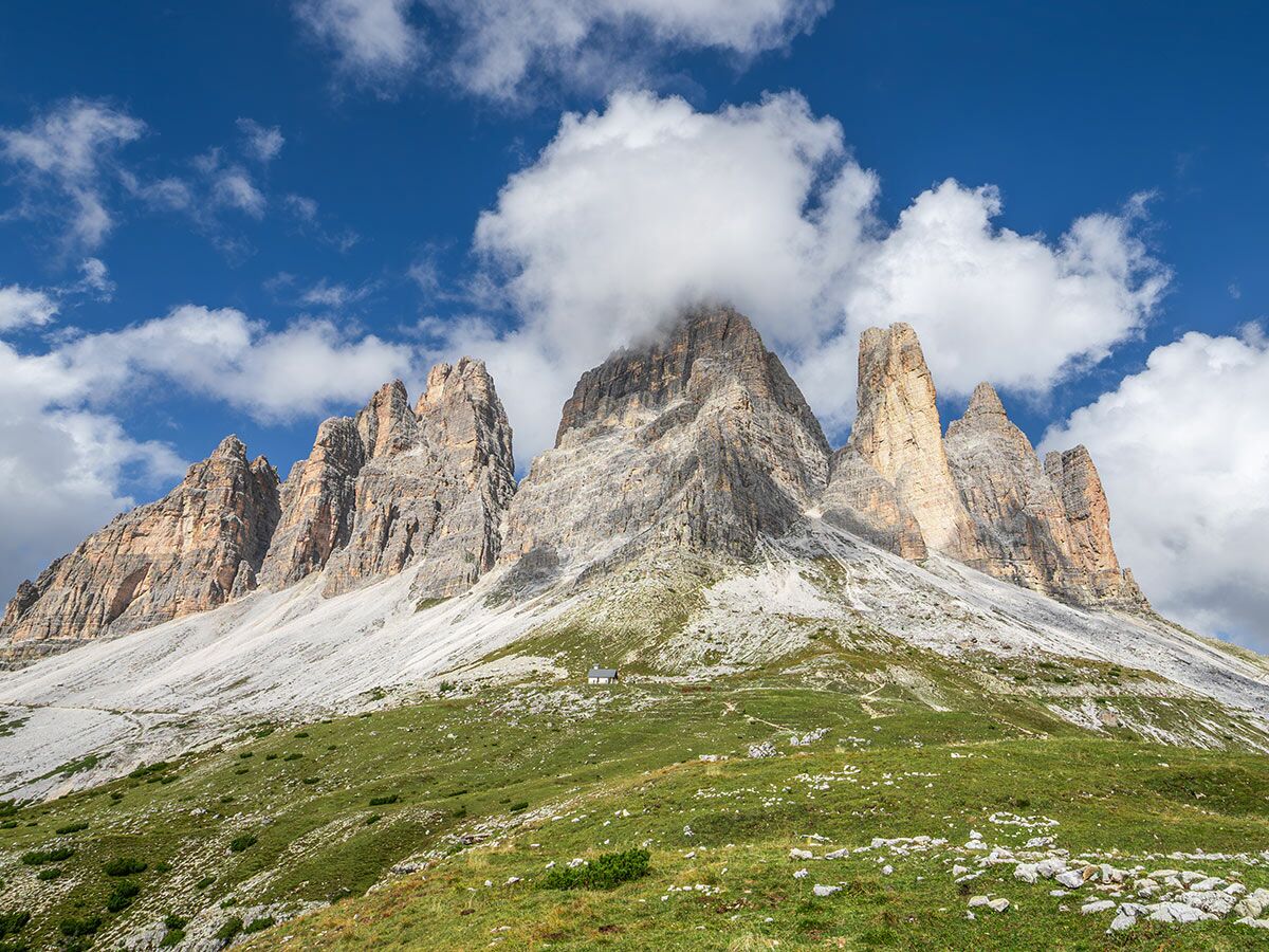 Majestätische Drei Zinnen in den Dolomiten unter blauem Himmel mit Wolken und grünen Hängen, kleines Gebäude sichtbar.