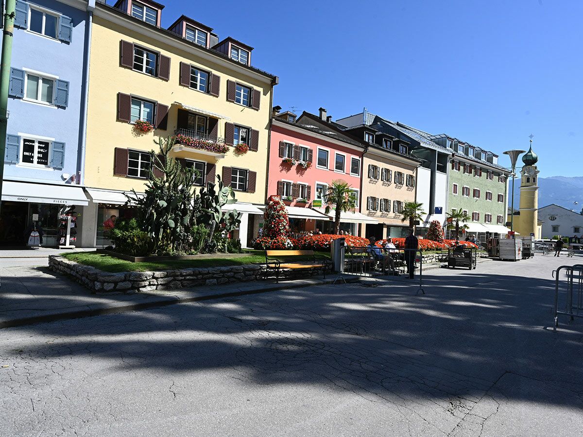 Bunter Stadtplatz in einer Alpenstadt mit farbigen Gebäuden, Blumen, Kakteen, Menschen und Bergkulisse unter blauem Himmel.
