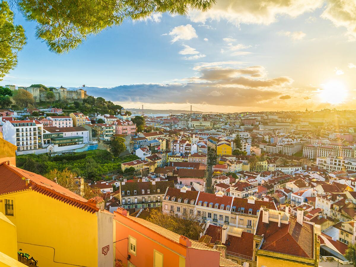 Lissabon Stadtpanorama bei Sonnenuntergang mit Castelo de São Jorge, roten Dächern und Tejo-Brücke im Hintergrund.