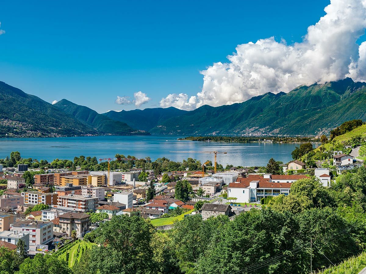 Panoramablick auf eine Alpenstadt an einem See mit grünen Bergen, blauem Himmel und weißen Wolken im Sommer.